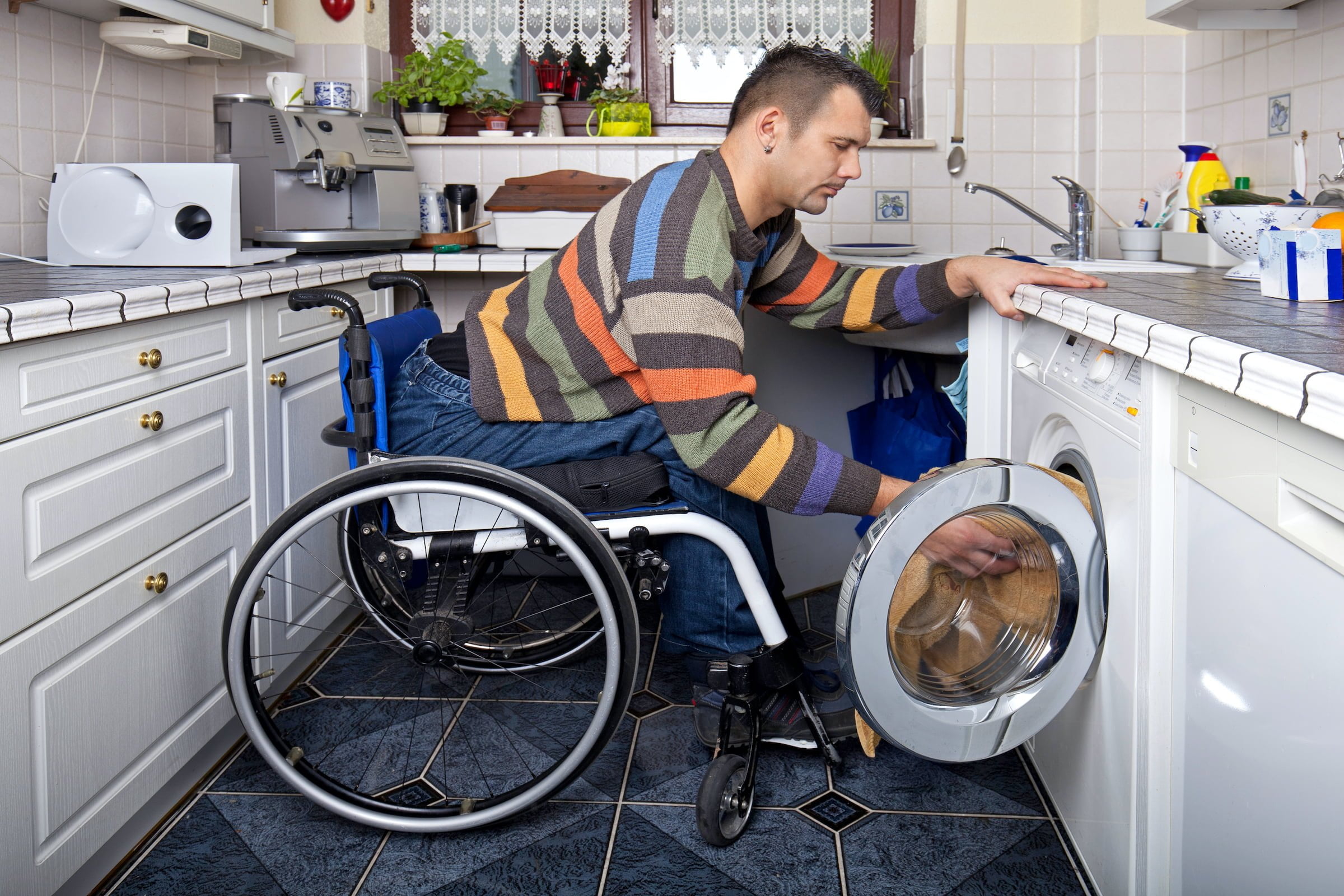 IL Skills Young white male sitting in a wheelchair leaning forward reaching laundry in a front load washer.