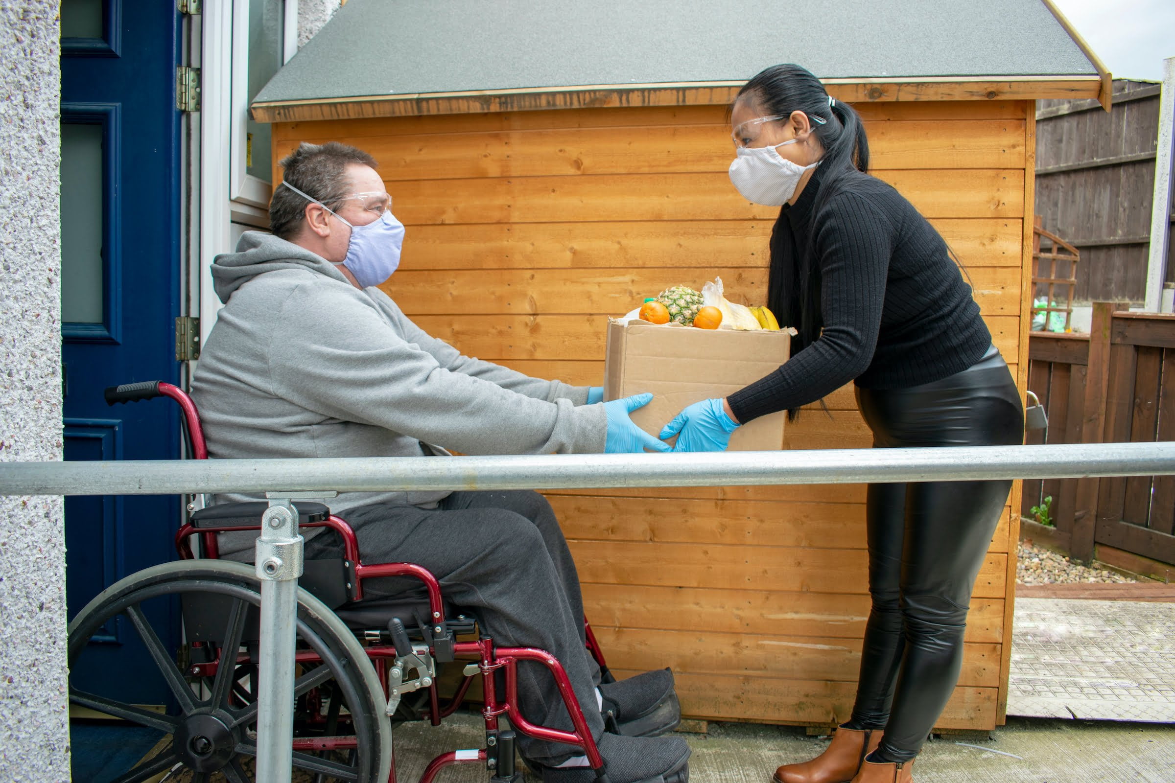 white male wearing a mask sits in a wheelchair while a woman hands him a box of groceries.