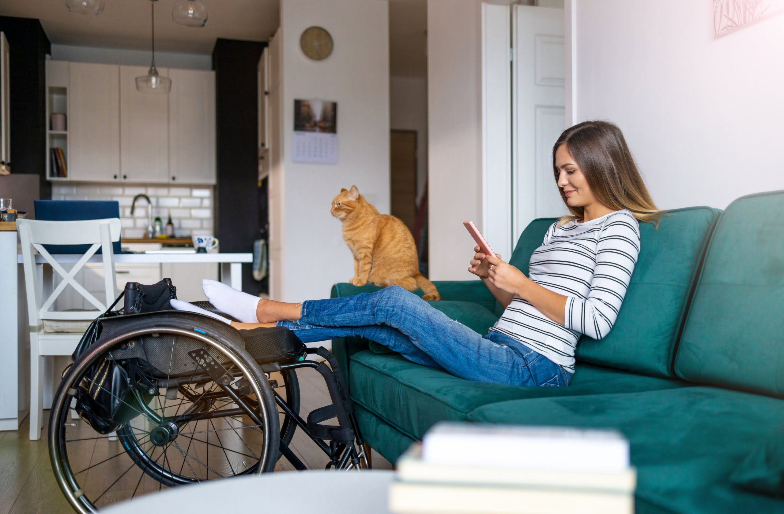 Young lady sits on a sofa looking at her phone. A cat sits on the armrest. Her legs are outstretched resting on a wheelchair.