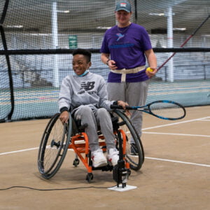 young black kid sitting in a sports wheelchair with a tennis racket in left hand. Another person standing just behind his left shoulder