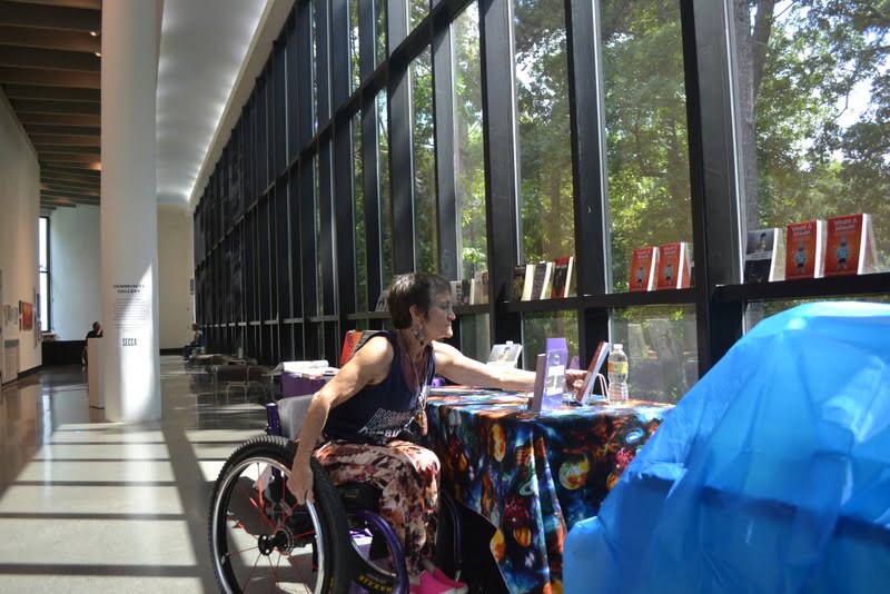 It is a picture of a mid age woman in a wheelchair reaching to put a book in place. The table has a table cloth on it that is colorful. The sun is coming from the windows lighting up the table full of book.