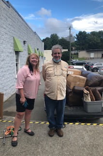 It is a picture of one of our consumer getting furniture for his home. It has a mid age woman with a pink shirt and black shorts. She is standing next to a old man with gray hair. He has on a tan shirt with blue jeans. Behind him is furniture that is going in his home.