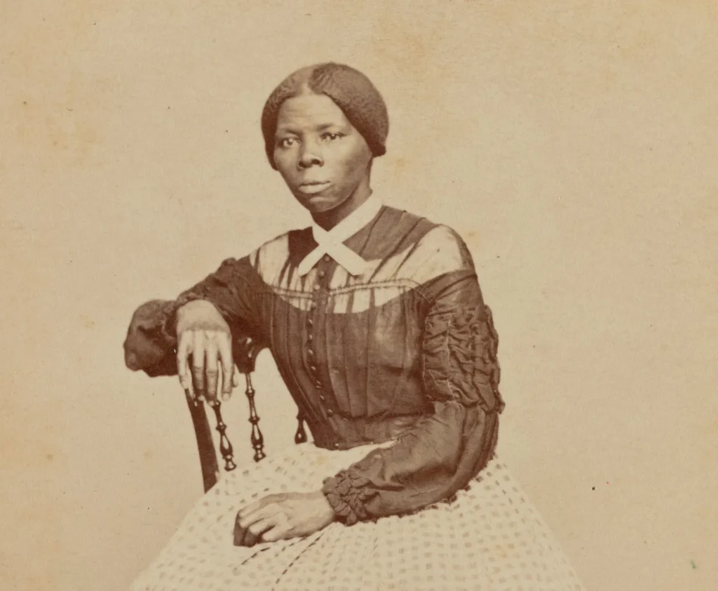 [A photo of Harriet Tubman. She has her hair tied back and wearing a dark dress with a white skirt. The photo has a brown tint from age.]