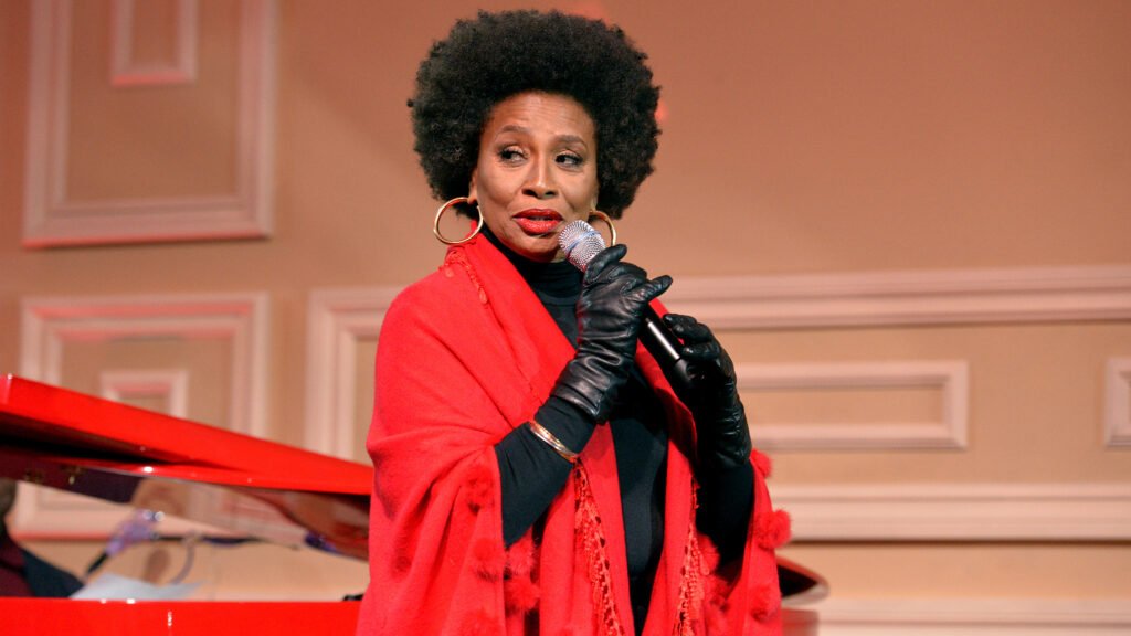 [A photo of Jenifer Lewis. She has an afro and large hoop earrings. She is wearing a red sweater and black turtleneck shirt with black gloves. She is holding a microphone to her mouth and turned to the side with her head turned toward the camera.]