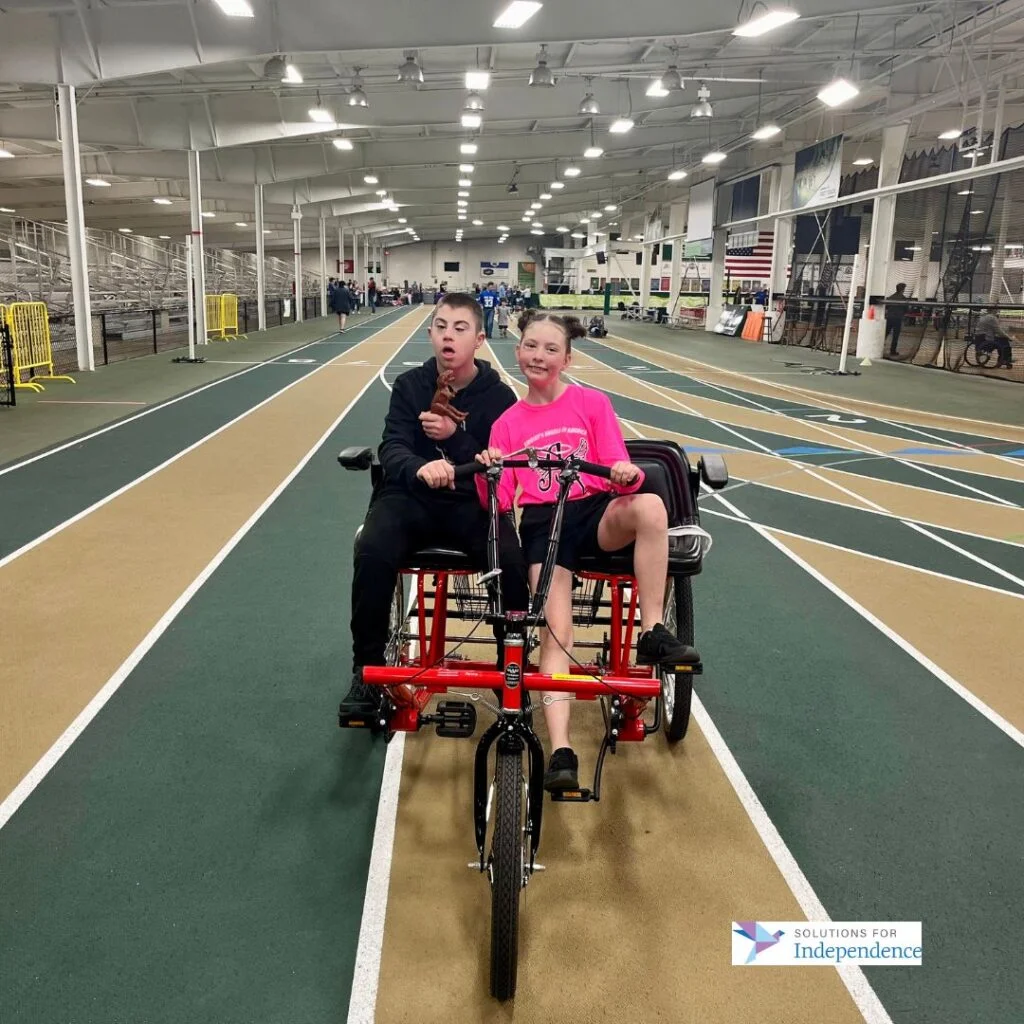 [A photo of two kids riding an adaptive bicycle. The boy on the left is wearing a black tracksuit. The girl on the right is wearing a pink t-shirt. They are smiling at the camera]