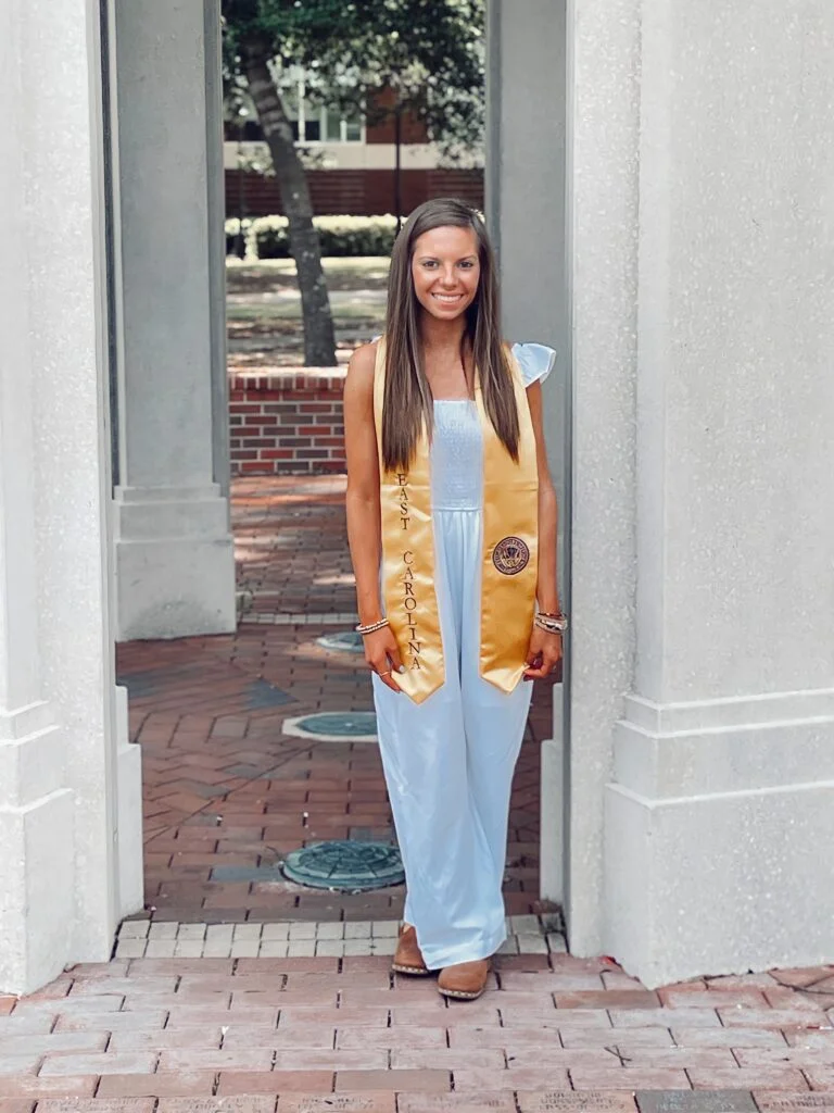 [A photo of Cameron Egan. She is standing next to a pillar and posing at the camera. She is wearing a light blue jumpsuit and a gold graduation sash. She is smiling at the camera.]
